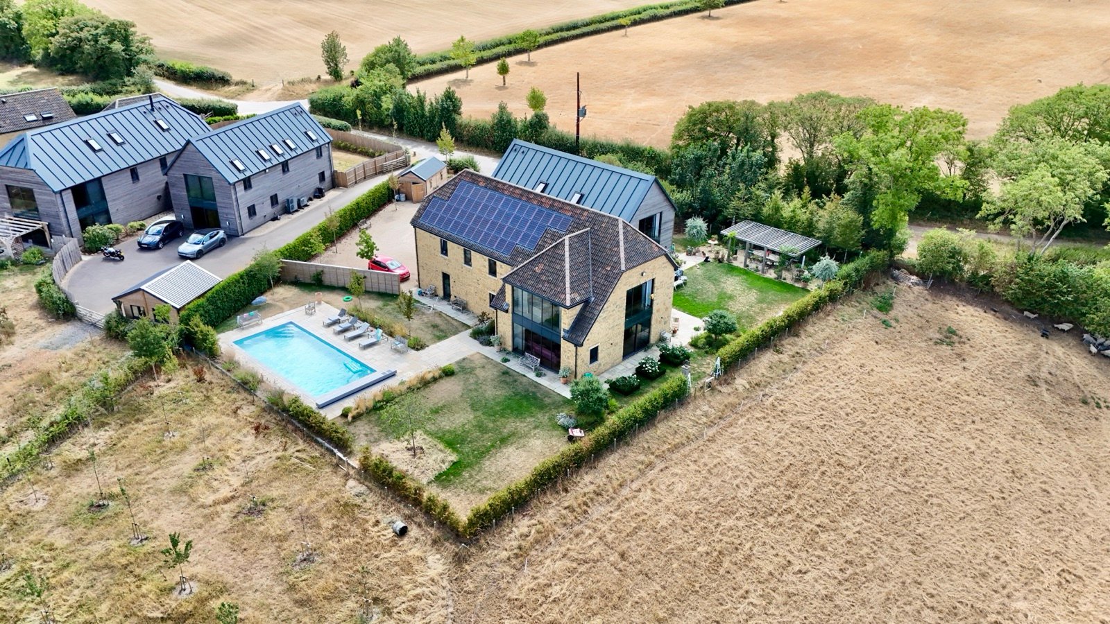 Modern stone house with solar panels, swimming pool and gardens bordered by open fields, aerial view